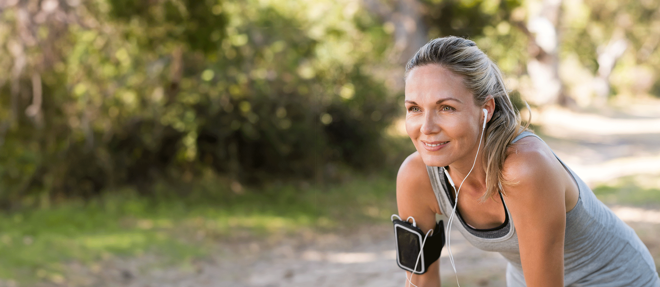 Sportieve vrouw met oordopjes en een smartphone-armband, genietend van een hardlooptraining in een groene, zonnige omgeving